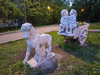 Horse statue and children is sitting on a cart. The scene is set in a park, with a path leading to...