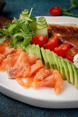 A balanced breakfast dish with fluffy omelette, sliced avocado, smoked salmon, cherry tomatoes, cucumber rolls, cream cheese and arugula on white plate