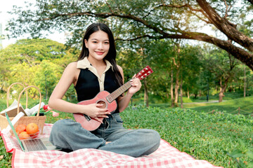 Portrait of smiling Asian beautiful girl plays ukulele while has a picnic in park. Young woman...