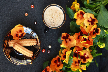 Top view of a cup of coffee and cookies on a dark textured surface, next to a bunch of orange pansies and decorative pearls.