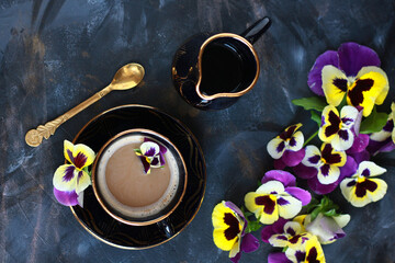 A cup of coffee with pansy flowers in a black and gold set on a dark textured background. Edible...