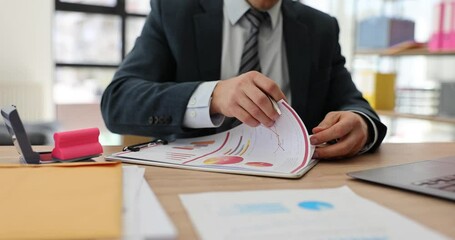 Businessman turns pages of financial reports with charts and graphs working at office desk. Focus on data analysis and strategic decision making - Powered by Adobe