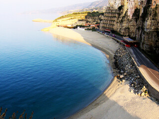 Stunning aerial view of the coastline in Tropea, Italy, showcasing the crystal-clear turquoise sea gently lapping against the golden sandy beach. The cliffs rise majestically above the shoreline
