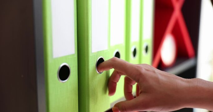 Office worker searches through files gently touching green ring binders arranged on shelf. Focus on organizing important documents in company office