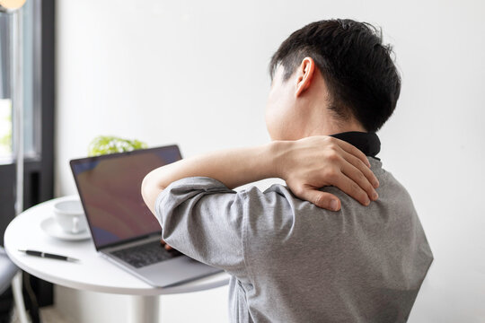Young Asian man experiencing neck and shoulder pain from working on a laptop, a common issue for modern office workers and a sign of prolonged desk work discomfort.