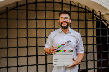 A hipster Latino man holding a clapperboard preparing to shoot a scene in a prison. Concept of cinema, movies, entertainment, and audiovisuals.