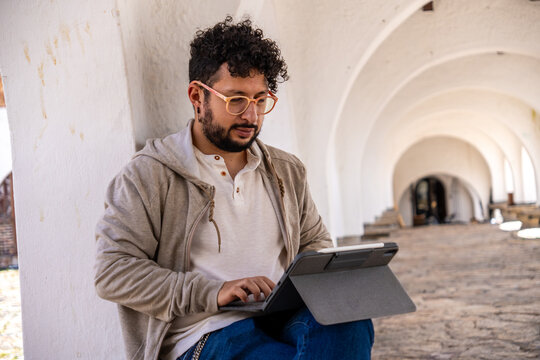 A Latino man wearing orange glasses and curly hair working remotely on a tablet in a castle in a colonial town in Colombia. Concepts of teleworking, connectivity, productivity, and travel.