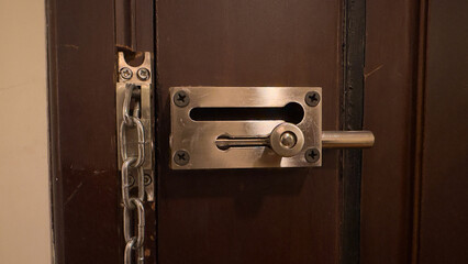 A close-up of a dark wooden door secured with a metal slide bolt and a hanging chain latch, both...
