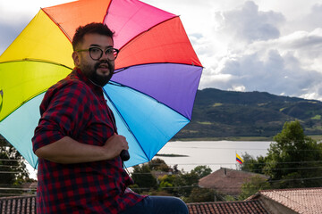 A hipster-style Latino man wearing glasses and holding a rainbow umbrella shows his support for the...