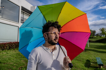 A curly-haired Latino man with orange glasses using a rainbow umbrella showing his support for the...