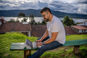 A Latino man working remotely with a tablet from a colonial village overlooking a lake and houses...