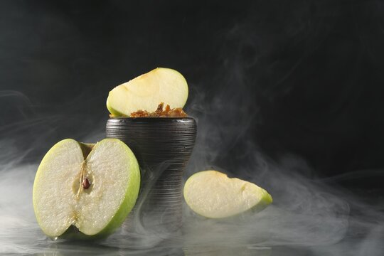 Hookah bowl with apple pieces and smoke on black table, closeup. Space for text