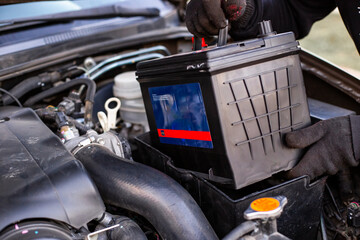 A man wearing black gloves installs a battery into a battery tray in the engine bay of a car. Replacing a battery in a car