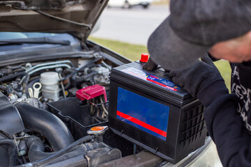 A man wearing gloves holds a battery pack over an engine bay, preparing to install it. Replacing a...