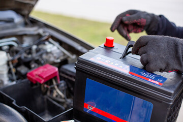 A man wearing gloves holds a battery pack over an engine bay, preparing to install it. Replacing a battery in a car