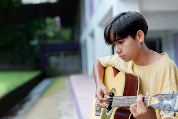 Teen boy playing acoustic guitar outdoors, wearing a yellow shirt, looking thoughtful. Creative youth expression and musical lifestyle in a casual setting.