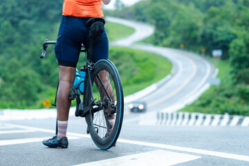 A cyclist in colorful gear stands on a winding road surrounded by greenery on a rainy day, ready to ride.