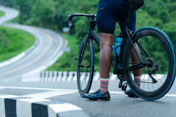 A cyclist stands with a road bike on a winding, rain-soaked road surrounded by lush greenery.