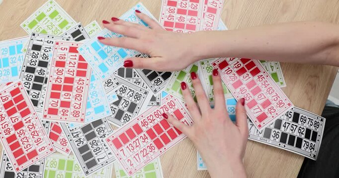 Woman hands sift through large spread of colorful bingo cards scattered on wooden table. Sorting activity involving numerous game sheets slow motion