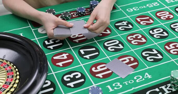 Woman distributes playing cards face down on green roulette table with numerous numbers and casino chips. Roulette wheel indicates game setting