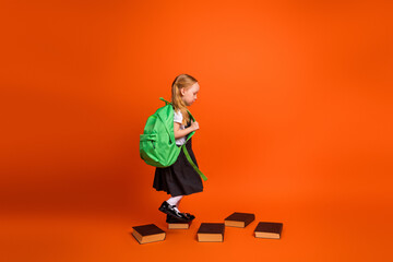 Adorable schoolgirl carrying a green backpack, walking on books on a bright orange background,...