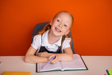 Naklejka premium Friendly schoolgirl with red hair smiling while studying in a classroom with a notebook and pencil on orange background