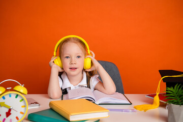Little girl wearing headphones studies at desk with school supplies on vibrant orange background