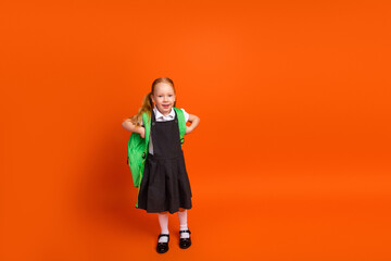 Cheerful schoolgirl with green backpack in uniform on orange background celebrating school and...