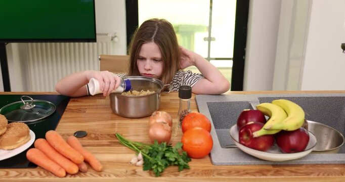Pensive preteen girl adds salt and pepper to dish into metal pot on wooden desk. Brown haired junior schoolgirl cooks dinner in lit home kitchen