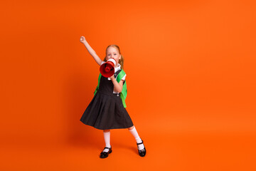 Adorable schoolgirl with backpack holding red megaphone against orange background, cheerful and...