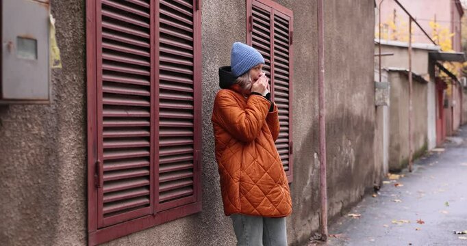 Woman in warm jacket and knitted hat blows runny nose standing near old building. Female suffers from cold symptoms and uses handkerchief on street