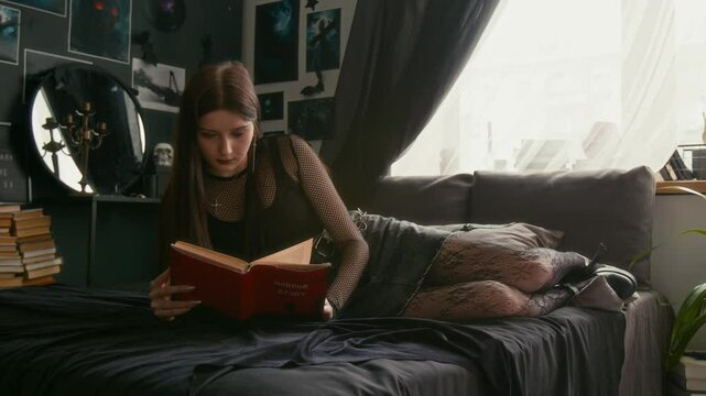 Young teenage goth girl lying on her bed and reading book in her dark gothic-themed bedroom with numerous wall posters