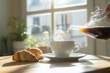 Pouring steaming coffee into cup with croissants near window in morning light