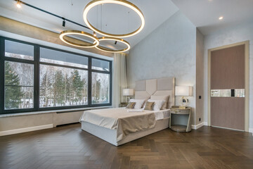 The bright and spacious bedroom has large windows overlooking the winter forest, modern furniture and elegant round lamps that create a cozy atmosphere.
