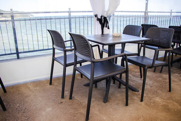 Black plastic chairs and table with small potted plant on a seaside terrace. Outdoor setting, ocean in background, white curtains and iron fence create a cozy coastal ambiance