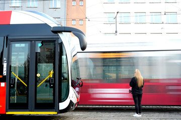 Modern red tram arrives at station while woman waits on platform. Motion blur emphasizes speed and...