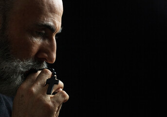 Elderly man with gray beard holding rosary beads in prayer against dark background. Weathered hands and contemplative profile create powerful spiritual moment of faith and devotion.
