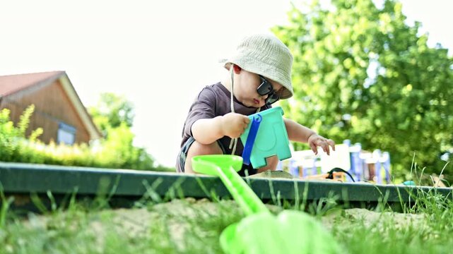 Toddler in a T-shirt and shorts. Little 2 year old boy playing in the sandbox on a playground outdoors. Toddler playing with a sand shovel. Creative activities for children in nature.