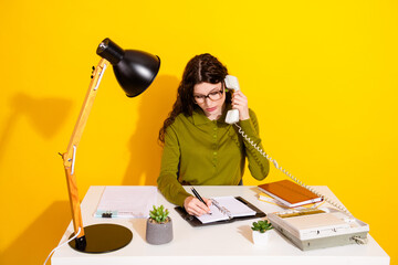 Young woman in green shirt using telephone while writing in notebook at desk with organized office supplies.