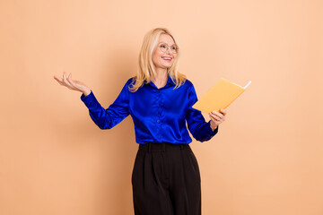 Elegant mature woman in blue blouse holding book against beige background showcasing confident and...