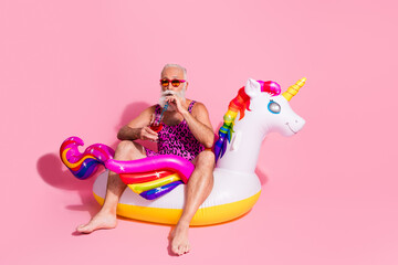 Playful senior man enjoying summer in pink leopard print swimsuit on inflatable unicorn against pink backdrop