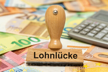 Ein Holzstempel ist beschriftet mit dem Aufdruck Lohnlücke und symbolisiert den Unterschied im Lohn zwischen Mann und Frau bzw. Ost und West