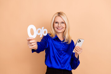 Mature woman in blue blouse holding smartphone and 0 percent sign, promoting discounts or financial...