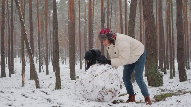 Two brothers apply a final, combined effort to push a heavy snowball up a small hill. This challenging teamwork is the last step in creating the base for their snowman in a winter forest.