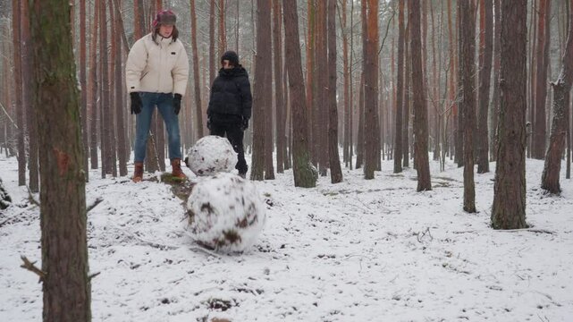 Two teen brothers push their large snowballs, letting them roll down a snowy hill in a winter forest. Then the happy siblings run down the slope, enjoying a fun and active outdoor game.