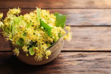 Beautiful linden flowers and green leaves in bowl on wooden table, closeup. Space for text