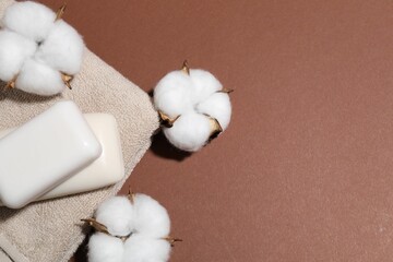Soap bars, cotton flowers and towel on brown background, flat lay. Space for text