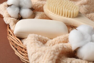 Soap bar, towel, brush and cotton flowers in wicker basket on brown background, closeup