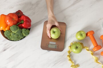 Woman weighting apple on kitchen scale at white marble table, top view