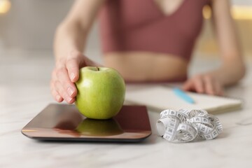 Woman weighting apple on kitchen scale at white marble table indoors, closeup